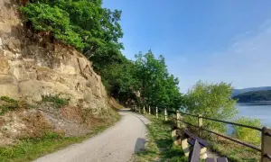 Ścieżka wzdłuż skalnego zbocza, z widokiem na jezioro i las. Krajobraz przypomina Eifel.