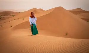 Kobieta w białej bluzce i zielonych spodniach stoi na piaszczystej wydmie. Czy Oman jest bezpieczny? Tak, to piękny kraj do zwiedzania.