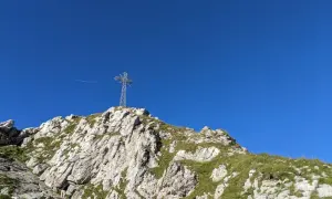 Na szczycie skalistego zbocza, porośniętego trawą, stoi metalowy krzyż. W tle błękitne niebo, a w oddali widać samolot. To widok z Giewontu, który przypomina, ile ma wysokości.