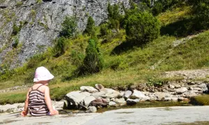 Dziecko w kapelusiku siedzi nad strumieniem w Małych Pieninach, otoczone zielenią i skalistym zboczem.