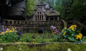 Kamienny domek w otoczeniu skał i zieleni, przypominający baśniową scenerię czeskiej Saksonii. Kobieta pochyla się nad kwiatami.