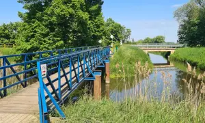 Błękitny mostek dla rowerzystów i pieszych nad kanałem na Żuławach Wiślanych. W tle widać kolejny most i bujną zieleń.