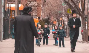 Dzieci w czapkach i zimowych kurtkach idą chodnikiem w Williamsburgu, trzymając paczki z przekąskami. Obok idzie mężczyzna w czarnym płaszczu i kapeluszu.
