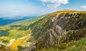 Widok na skaliste zbocze i zieloną dolinę z jeziorami. Śnieżne kotły ukazują swoją wysokość w majestatycznym krajobrazie.