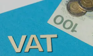 Drewniane litery VAT na niebieskim tle, obok banknot 100 zł i monety. Rozważanie vat należny a naliczony.
