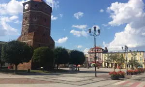 Wieża zegarowa w Barcinie na tle błękitnego nieba z chmurami, plac miejski z latarniami i budynkami.