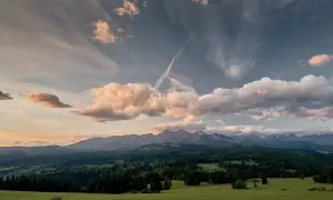 Widok na Tatry z zielonej łąki, zalesione wzgórza i chmury na niebie.