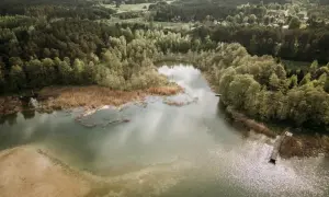 Widok z lotu ptaka na spokojne jezioro otoczone lasem, z piaszczystym brzegiem i drewnianym pomostem.