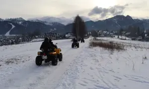 Ludzie na quadach jadą przez ośnieżony teren z widokiem na góry i zabudowania.
