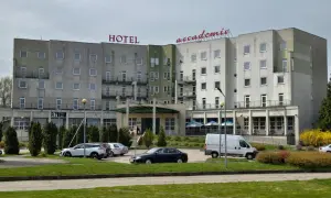 Hotel Accademia w Przemyślu z czerwonym napisem na dachu, parking z samochodami i białą furgonetką.