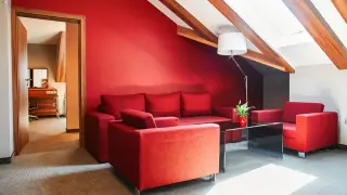 Czerwona kanapa i fotele w przytulnym pokoju hotelowym. W tle widać drzwi do kolejnego pomieszczenia.