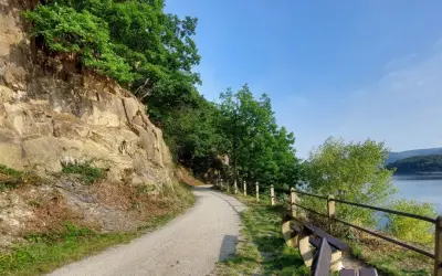 Ścieżka wzdłuż skalnego zbocza, z widokiem na jezioro i las. Krajobraz przypomina Eifel.
