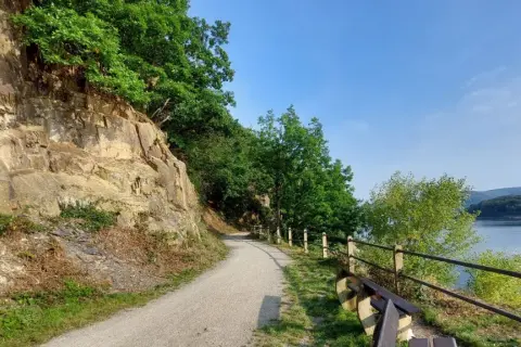Ścieżka wzdłuż skalnego zbocza, z widokiem na jezioro i las. Krajobraz przypomina Eifel.