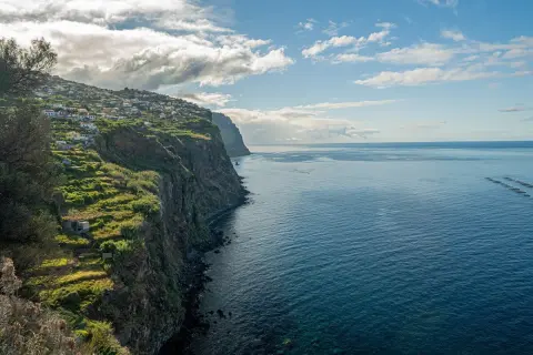 Widok na klif z domkami na zboczu i oceanem. Zastanawiasz się, ile się leci na Maderę, patrząc na ten krajobraz?