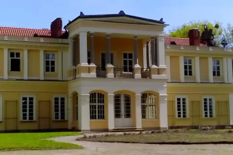 Pałac w Waplewie z kolumnowym portykiem i żółtą fasadą, otoczony zielenią.