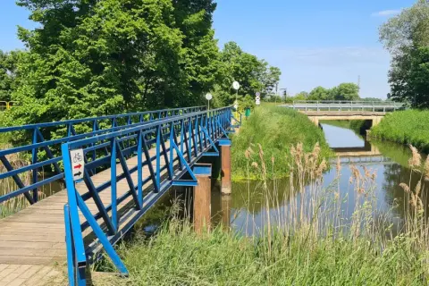 Błękitny mostek dla rowerzystów i pieszych nad kanałem na Żuławach Wiślanych. W tle widać kolejny most i bujną zieleń.