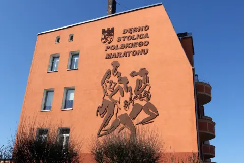 Pomarańczowa ściana budynku z płaskorzeźbą biegaczy i napisem "DĘBNO STOLICA POLSKIEGO MARATONU".