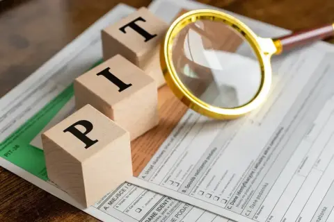 Drewniane klocki z literami "PIT" leżą na formularzu podatkowym. Lupa bada szczegóły.