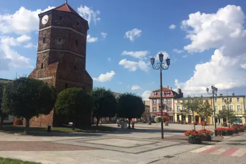 Wieża zegarowa w Barcinie na tle błękitnego nieba z chmurami, plac miejski z latarniami i budynkami.