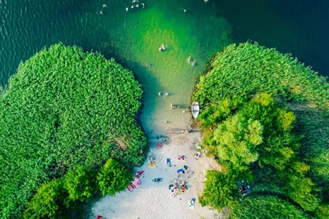 Letnie orzeźwienie nad jednym z najpiękniejszych jezior w Polsce. Ludzie kąpią się, plażują, a łódka czeka przy brzegu.