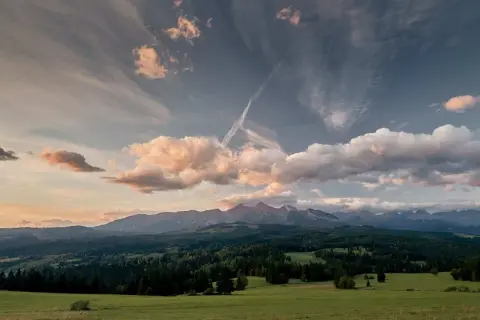 Widok na Tatry z zielonej łąki, zalesione wzgórza i chmury na niebie.