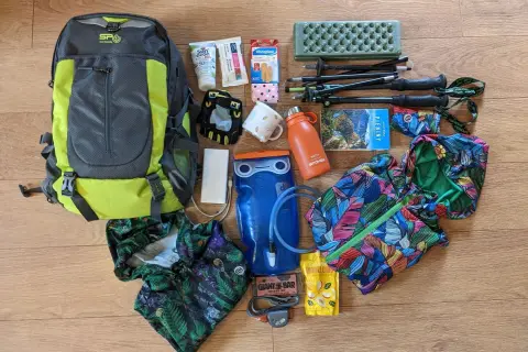 Plecak, kijki trekkingowe, bidon, apteczka i inne akcesoria turystyczne ułożone na drewnianym tle.