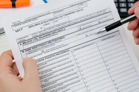 Dłoń z długopisem wypełnia deklarację VAT-7, dokument podatkowy z polami do wpisania danych.