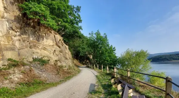 Ścieżka wzdłuż skalnego zbocza, z widokiem na jezioro i las. Krajobraz przypomina Eifel.