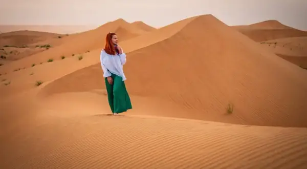 Kobieta w białej bluzce i zielonych spodniach stoi na piaszczystej wydmie. Czy Oman jest bezpieczny? Tak, to piękny kraj do zwiedzania.