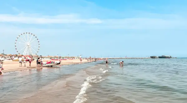 Słoneczny dzień na plaży w Rimini. Ludzie spacerują, bawią się w wodzie, a w oddali widać diabelski młyn. Idealne miejsce na wakacje!