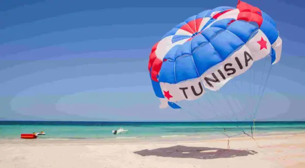 Kolorowy spadochron z napisem "TUNISIA" unosi się nad plażą, zapowiadając wspaniałe dżerba loty. W tle turkusowe morze i piaszczysta plaża.