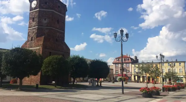 Wieża zegarowa w Barcinie na tle błękitnego nieba z chmurami, plac miejski z latarniami i budynkami.
