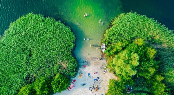Letnie orzeźwienie nad jednym z najpiękniejszych jezior w Polsce. Ludzie kąpią się, plażują, a łódka czeka przy brzegu.