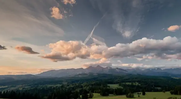 Widok na Tatry z zielonej łąki, zalesione wzgórza i chmury na niebie.