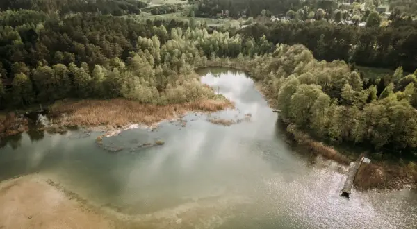 Widok z lotu ptaka na spokojne jezioro otoczone lasem, z piaszczystym brzegiem i drewnianym pomostem.