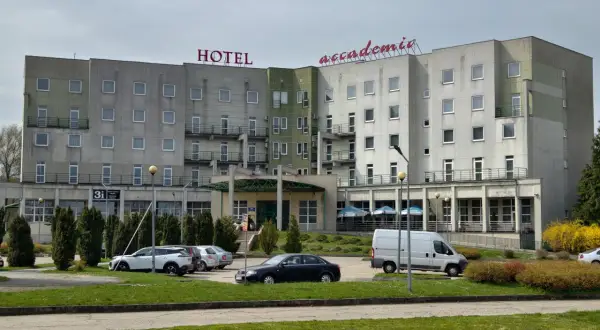 Hotel Accademia w Przemyślu z czerwonym napisem na dachu, parking z samochodami i białą furgonetką.