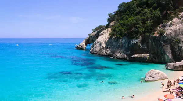 Rajska plaża Cala Goloritze z turkusową wodą, skalistym klifem i łukiem skalnym. Ludzie kąpią się i odpoczywają na piasku.