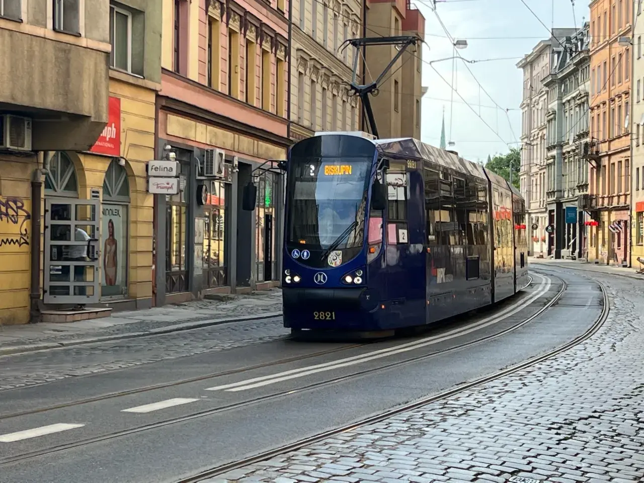 Niebieski tramwaj z napisem "Biskupin" na wyświetlaczu, jadący po brukowanej ulicy w mieście.