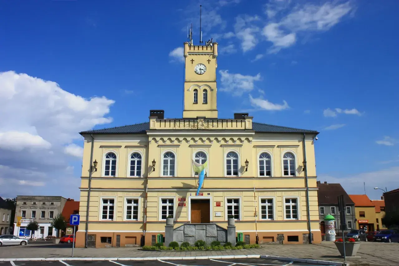 Żółty budynek ratusza z wieżą zegarową i flagą na tle błękitnego nieba.
