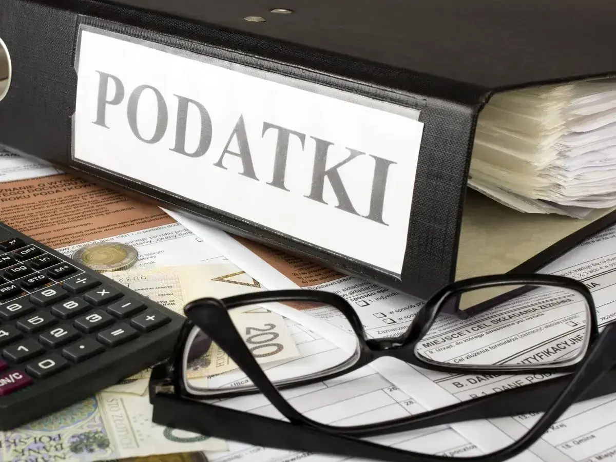Segregator z napisem "PODATKI", okulary, kalkulator i pieniądze na dokumentach. Czas na rozliczenie PIT-2.