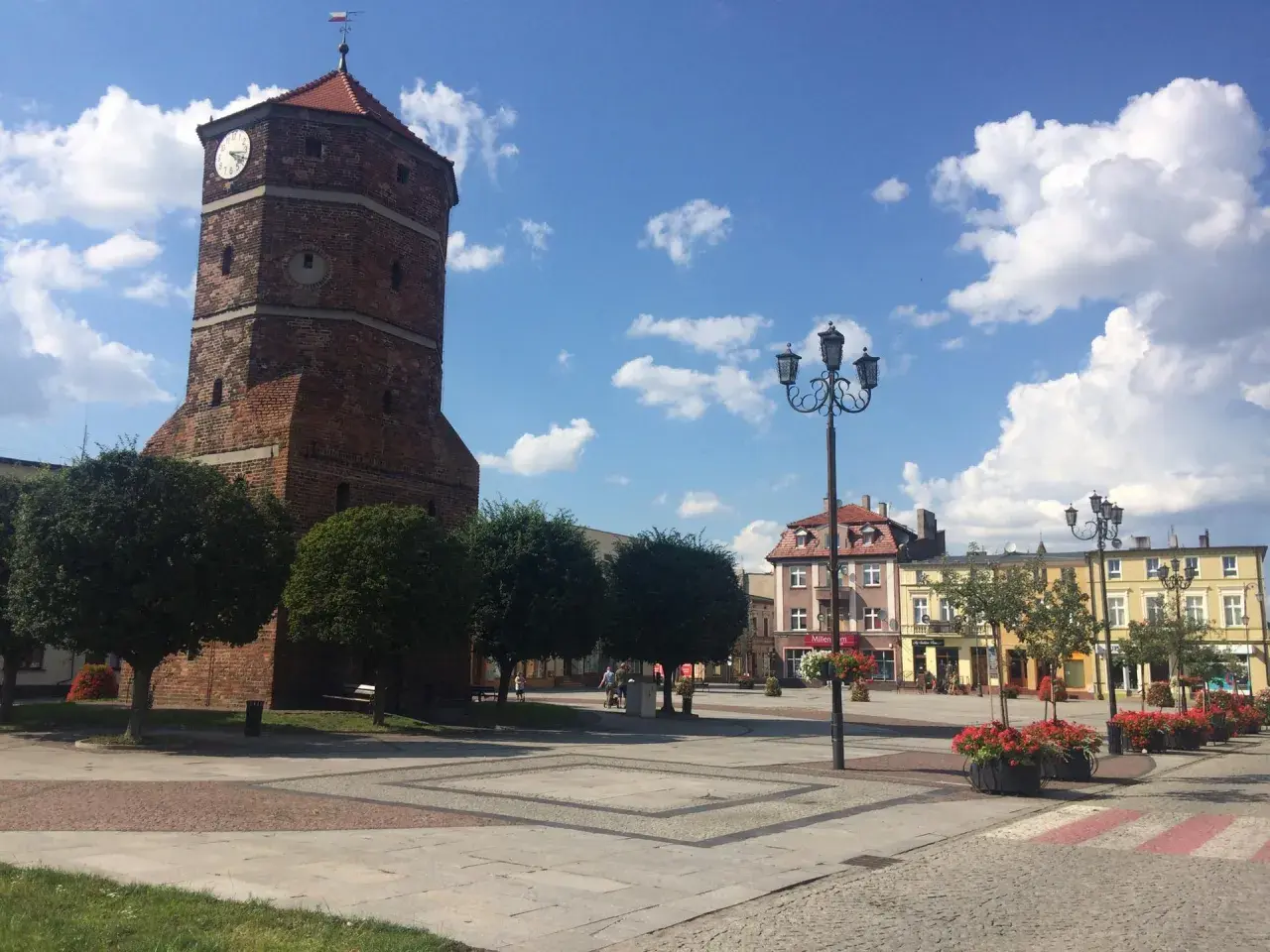 Wieża zegarowa w Barcinie na tle błękitnego nieba z chmurami, plac miejski z latarniami i budynkami.