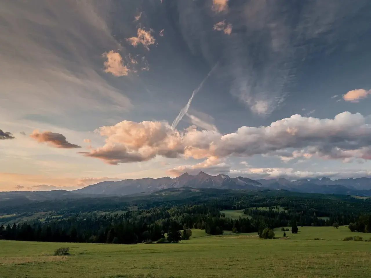 Widok na Tatry z zielonej łąki, zalesione wzgórza i chmury na niebie.