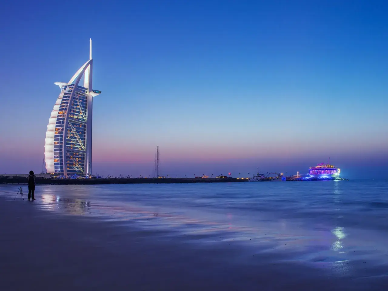 Wieżowiec Burdż al-Arab i oświetlony statek na tle nocnego nieba nad Zatoką Perską.