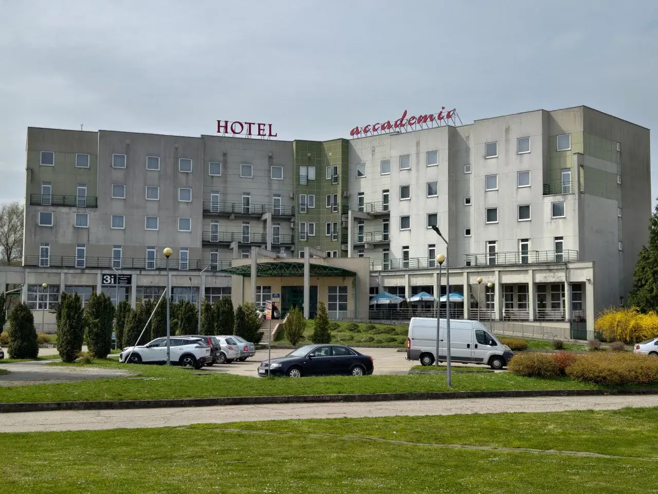 Hotel Accademia w Przemyślu z czerwonym napisem na dachu, parking z samochodami i białą furgonetką.
