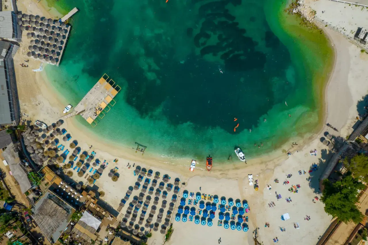 Rajskie **Albania wakacje**! Widok z lotu ptaka na turkusowe morze, piaszczystą plażę z leżakami i parasolami, drewniane molo i łodzie.