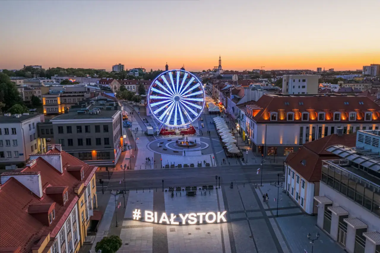 Widok z góry na plac z podświetlanym napisem #BIAŁYSTOK i diabelskim młynem o zachodzie słońca.
