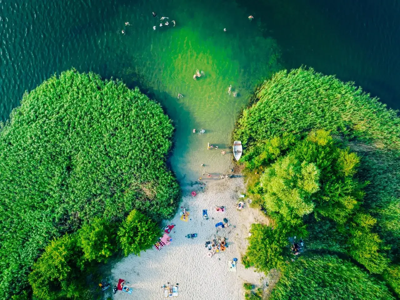 Letnie orzeźwienie nad jednym z najpiękniejszych jezior w Polsce. Ludzie kąpią się, plażują, a łódka czeka przy brzegu.
