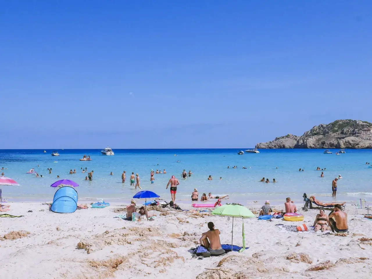 Słoneczna plaża Cala Agulla tętni życiem. Ludzie kąpią się w krystalicznie czystej wodzie, opalają pod parasolami i relaksują na piasku.