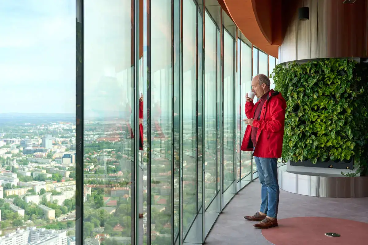 Mężczyzna w czerwonej kurtce pije kawę, podziwiając panoramę Wrocławia. Sprawdź sky tower wrocław cennik, by poznać ceny wejścia.