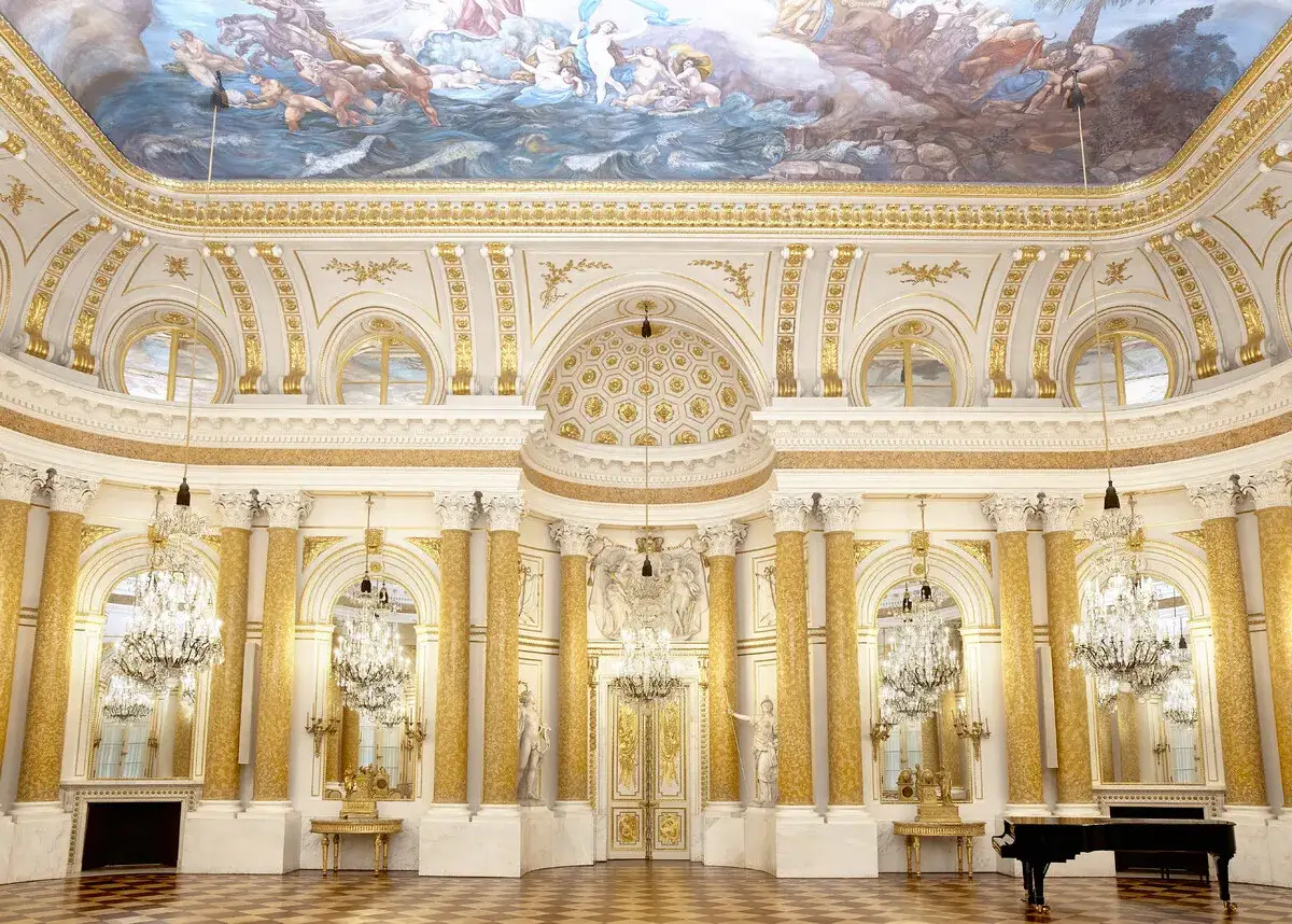 Wnętrze Zamku Królewskiego w Warszawie: bogato zdobiona sala balowa z malowidłami na suficie, kryształowymi żyrandolami i złotymi zdobieniami.
